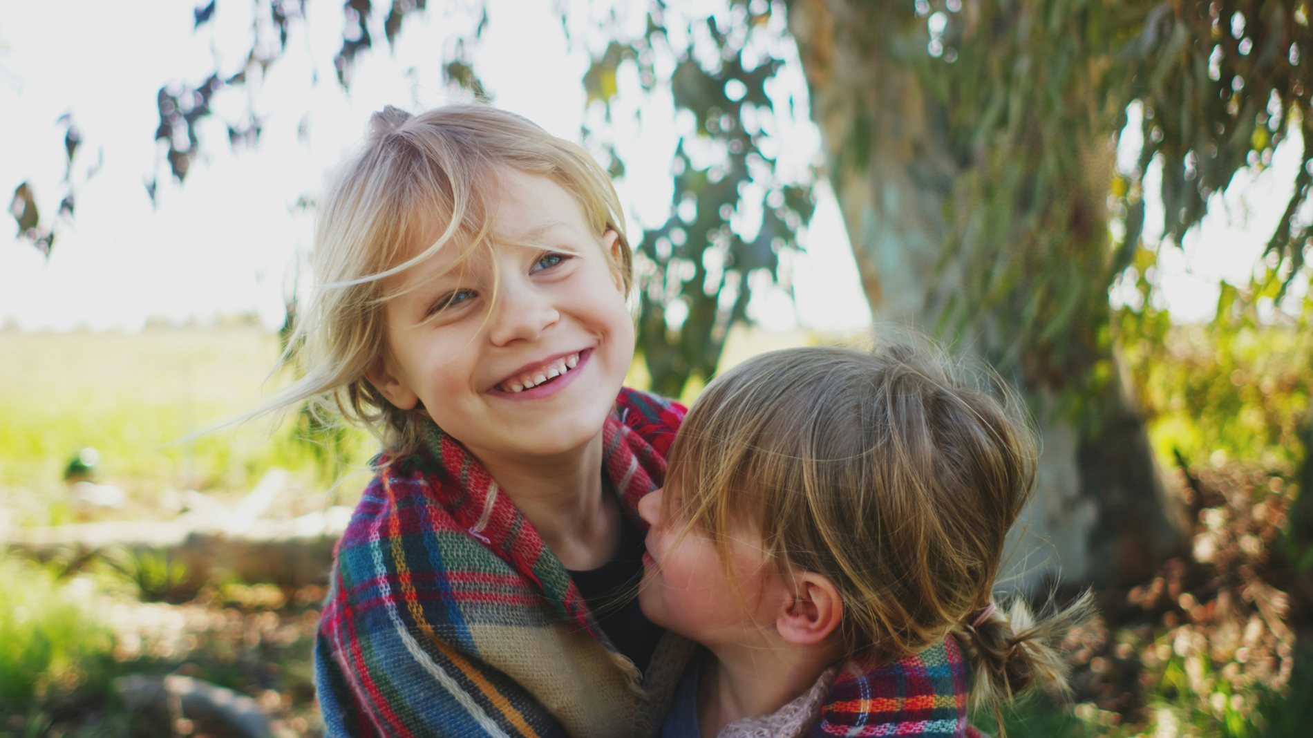 Two young children wrapped together in a blanket under a tree